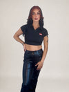 A woman modeling a black short-sleeve ideal apparel cropped polo shirt with a small red and white logo patch on the left chest, paired with shiny black pants, against a plain light gray studio background.