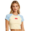 A woman modeling a short-sleeve ideal apparel crop top with a cream body and light blue raglan sleeves, featuring a red rectangular ideal apparel chest logo that reads 'ideal?'.