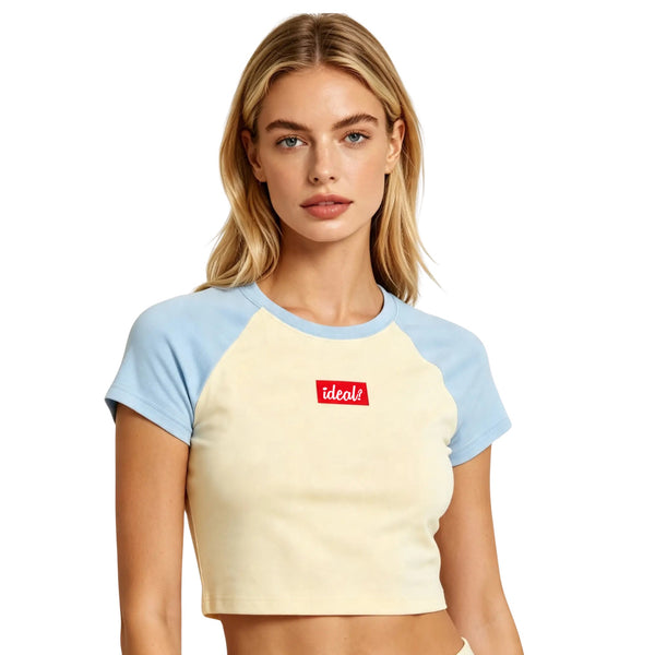A woman modeling a short-sleeve ideal apparel crop top with a cream body and light blue raglan sleeves, featuring a red rectangular ideal apparel chest logo that reads 'ideal?'.