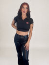 A woman modeling a black short-sleeve ideal apparel collared crop top with a small red and white logo patch on the left chest, paired with black faux-leather pants, against a plain light gray studio background.